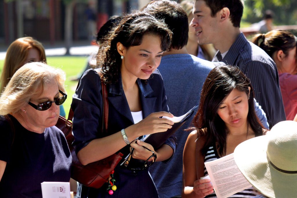 Diverse group of people outdoors, some reading documents and looking at papers.
