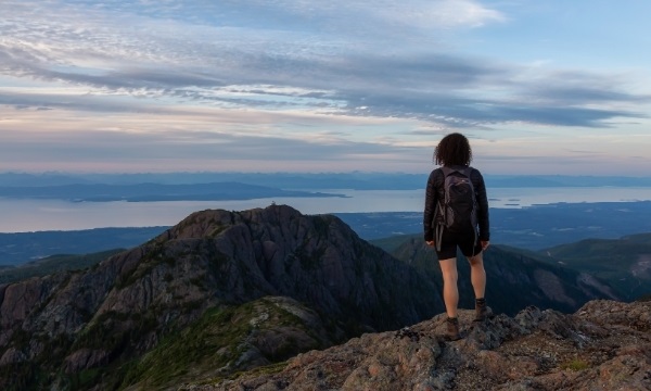 Hiker with backpack on mountain peak overlooks ocean and distant islands at sunset.
