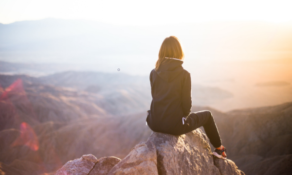 Woman in hoodie sitting on a rock overlooking a desert landscape at sunset.