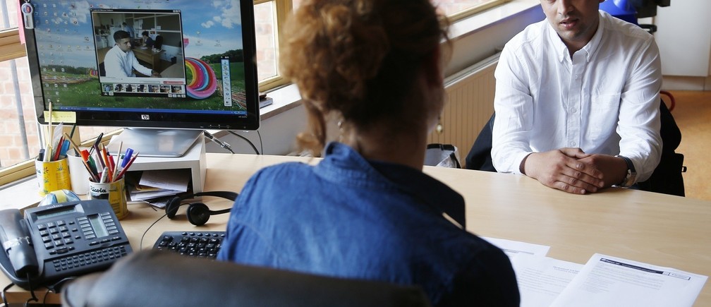 Two people in an office with a monitor displaying a video call, desk with phone and keyboard.