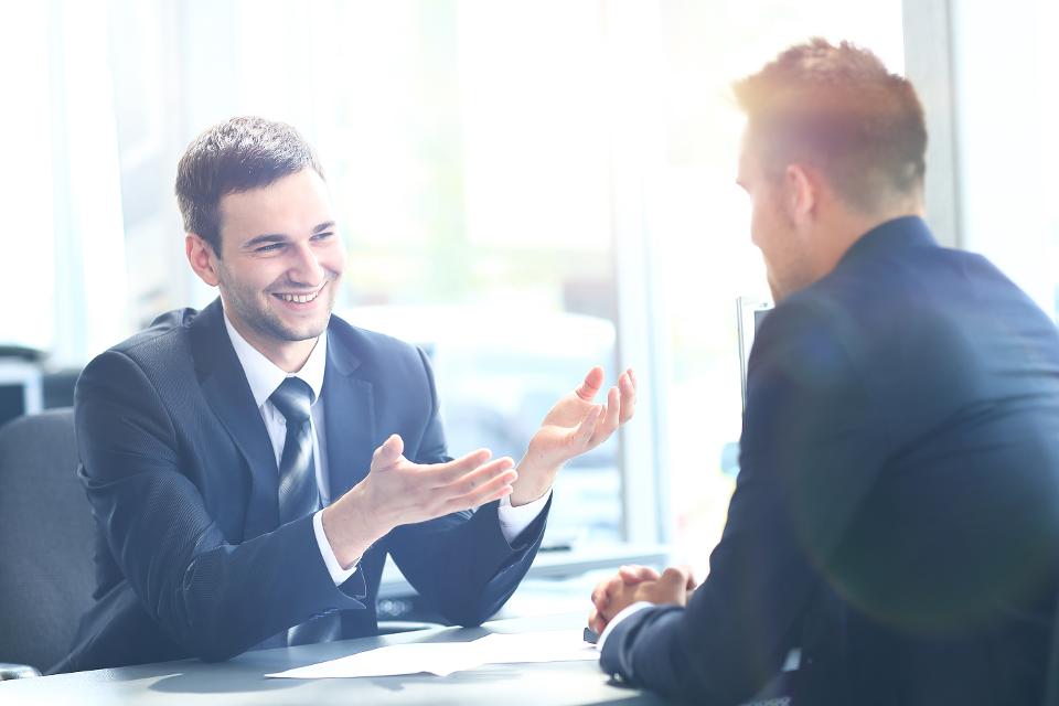 Two businessmen in suits talking at a table in a bright office, one gesturing.