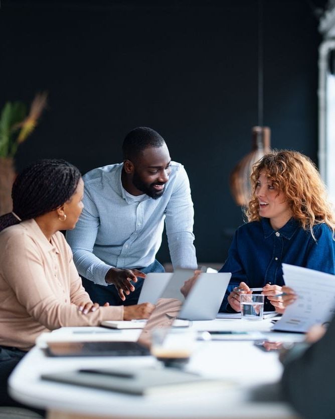 Diverse professionals in a meeting, collaborating around a table with laptops and documents.
