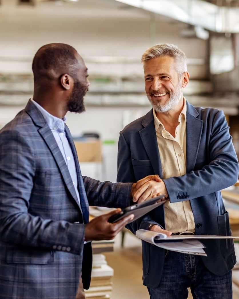Two men in suits shake hands, one holding a tablet, in a workshop.