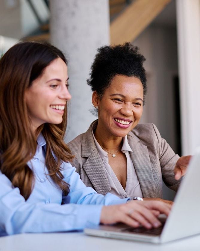Two smiling women, one with long brown hair and the other with short curly black hair, working together on a laptop.