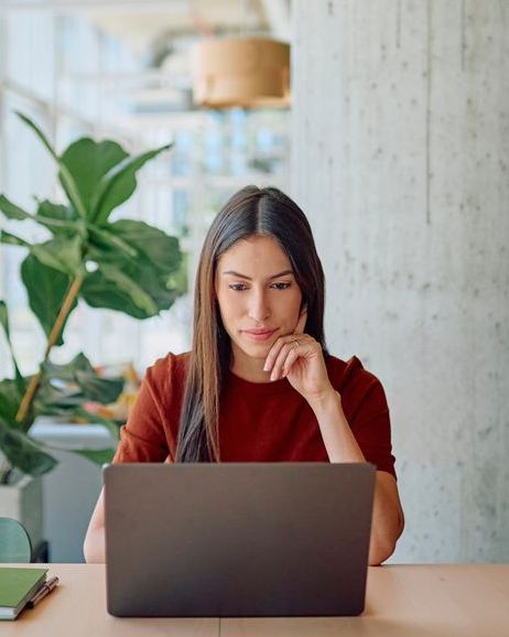 Woman with long brown hair wearing a red shirt, focused on her laptop.