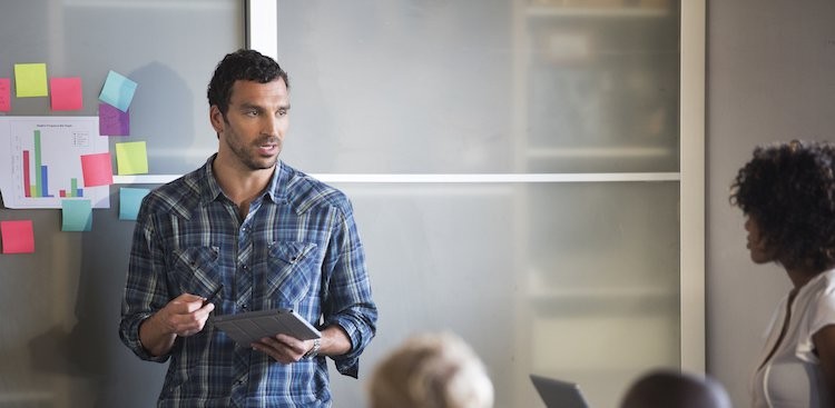 Man in plaid shirt presents with tablet in front of a whiteboard with sticky notes and a chart.