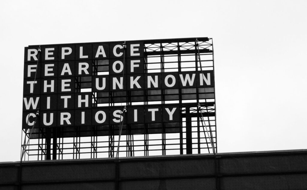 Black and white sign with text "REPLACE FEAR OF THE UNKNOWN WITH CURIOSITY" against a sky.