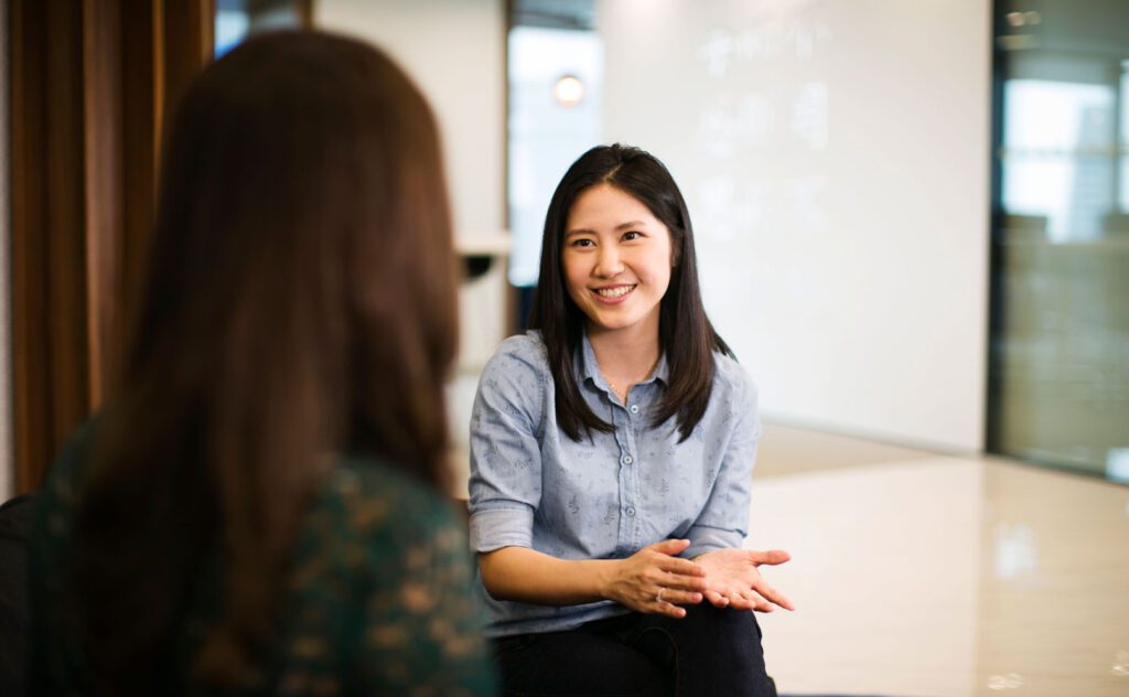 Two women in an office, one smiling and gesturing while talking to another woman.