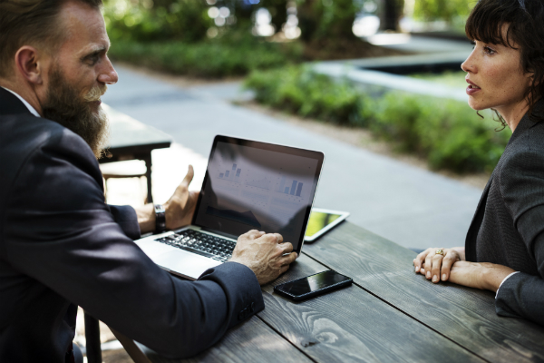 A man with a beard and a woman in suits discuss a laptop with charts outdoors.