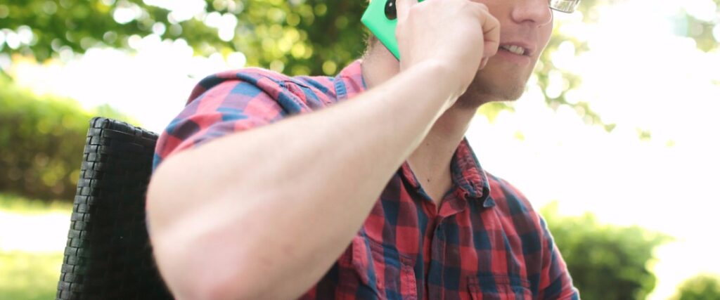 Man in red plaid shirt talking on a bright green smartphone outdoors, with bokeh background.